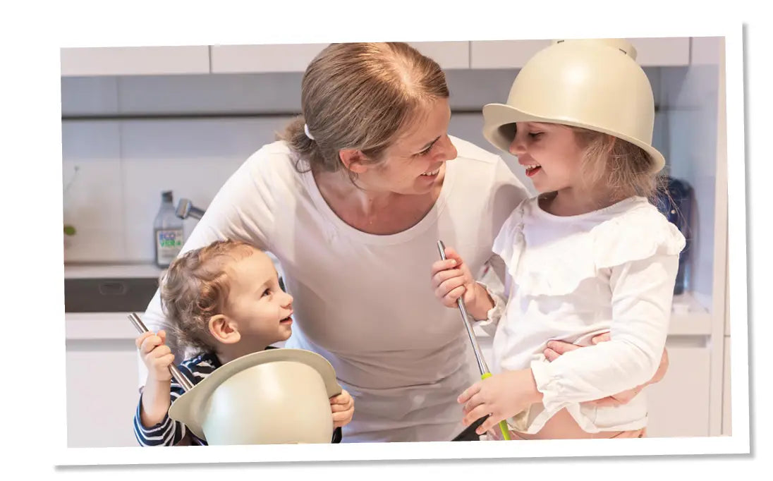 Bild einer Familie die in der Küche steht, ein Kind hat eine Rührschüssel auf dem Kopf, das andere Kind rührt in einer Rührschüssel, alle lachen