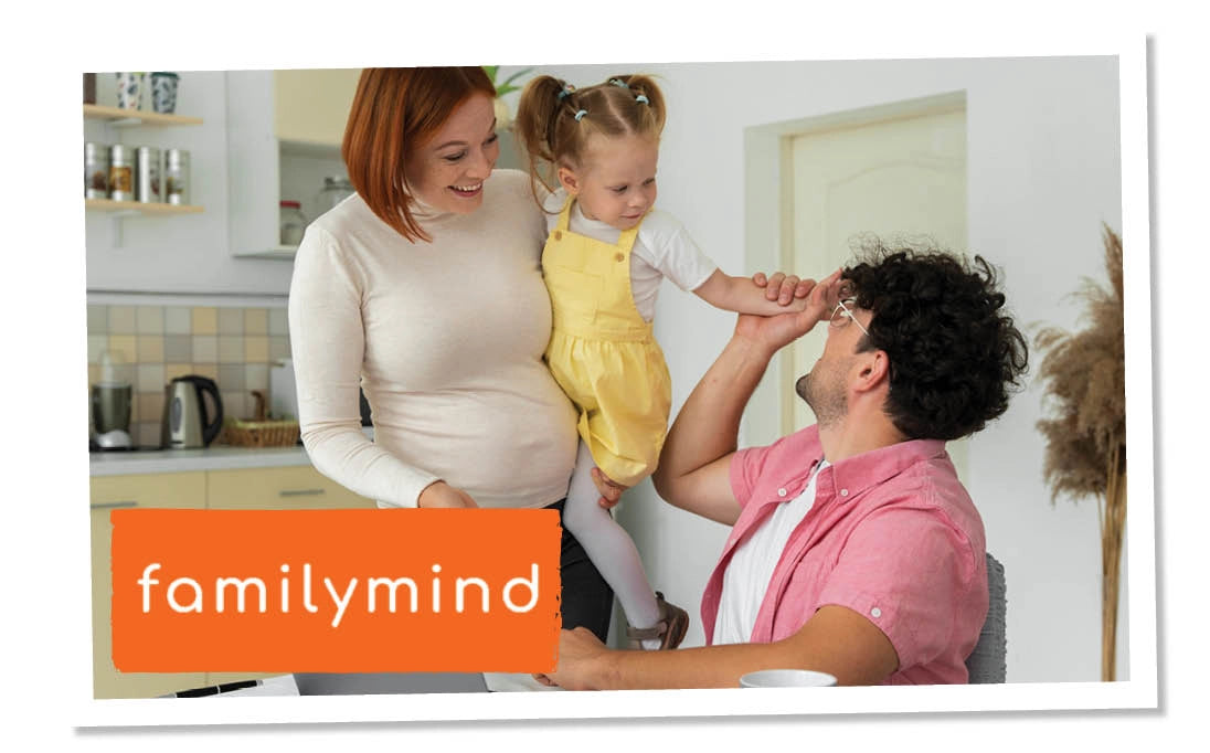 Schwangere mit Kind im Arm, Mann sitzt am Tisch und interagiert mit Kind